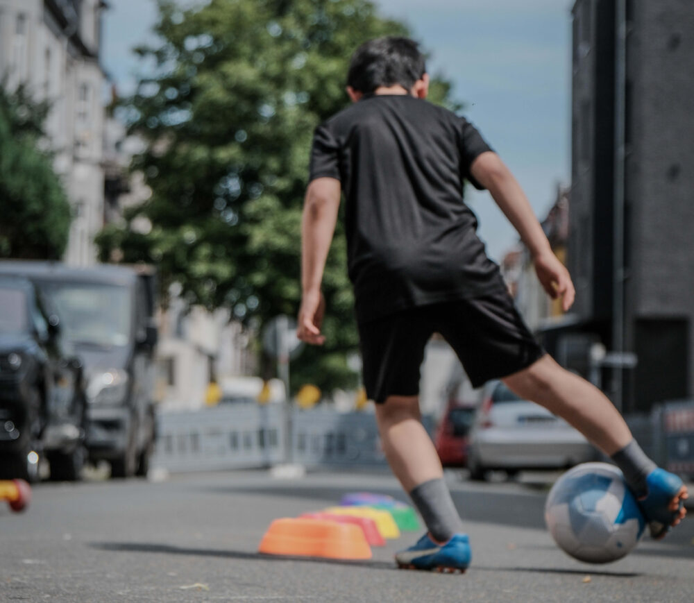 Ein Kind ist von hinten zu sehen wie es mit einem Fußball spielt. Es trägt ein schwarzes Trickot und Fußballschuhe