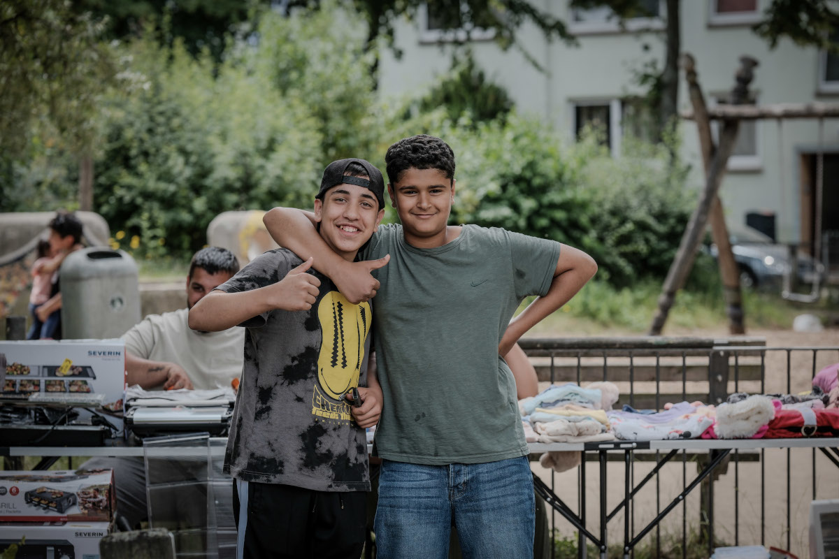 Zwei junge Menschen umarmen sich und schauen in die Kamera. Eine Person hält die Hand mit Daumen hoch. Im Hintergrund sind Bäume, Sträucher und Häuser.