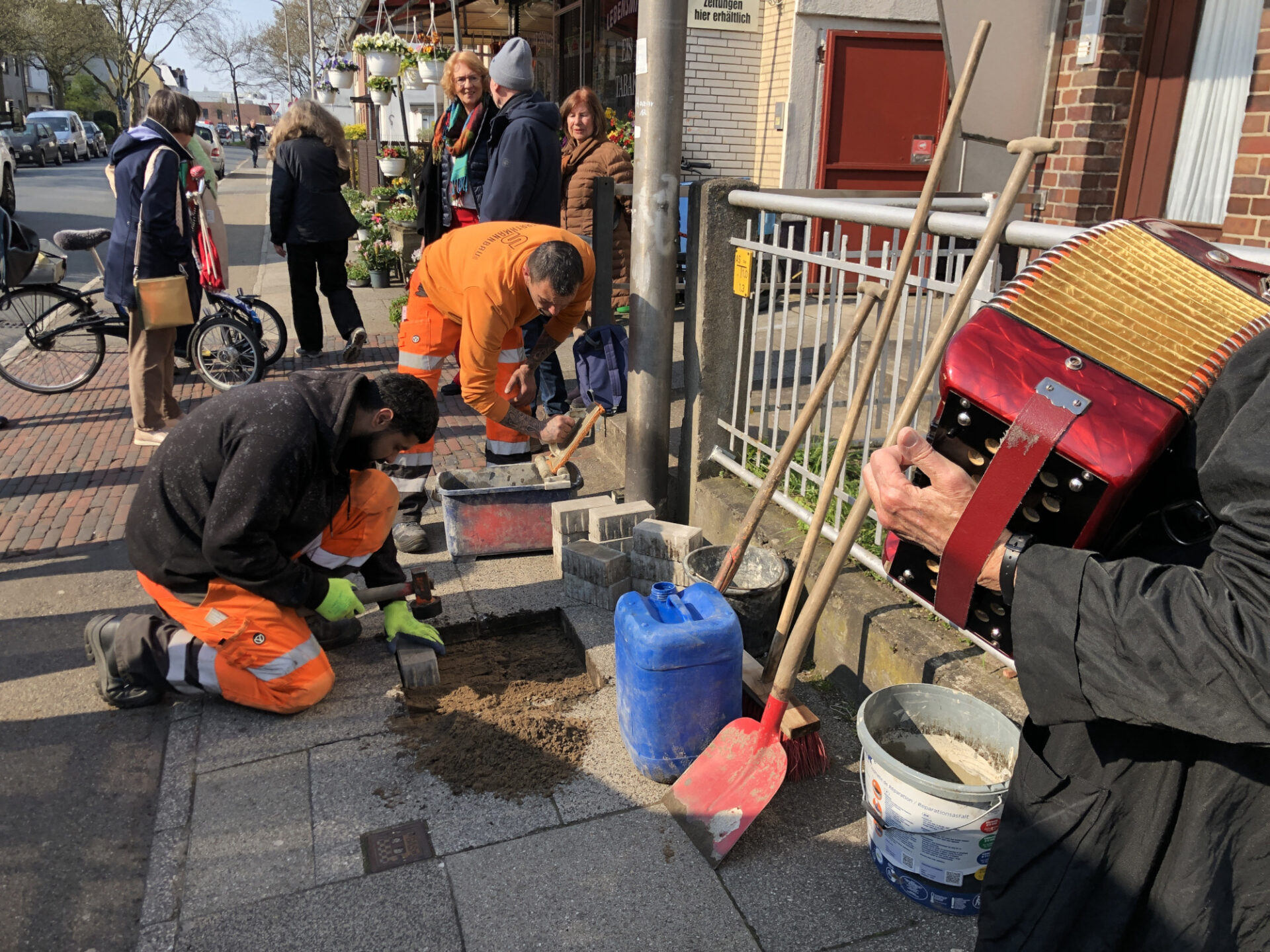 Zwei Personen in Arbeitskleidung verlegen auf dem Gehweg einen Stolperstein. Menschen stehen wartend daneben. Ein Akkordeon ist im anschnittrechts im bild zu sehen.