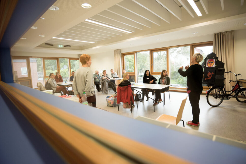 Durch ein schmales Fenster kann in einen Raum geschut werden, in dem Menschen um Tische sitzen und stehen.