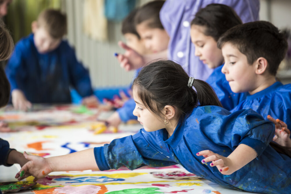 Kinder malen mit bunten Farben auf ein Papier, dass auf einem Tisch liegt.