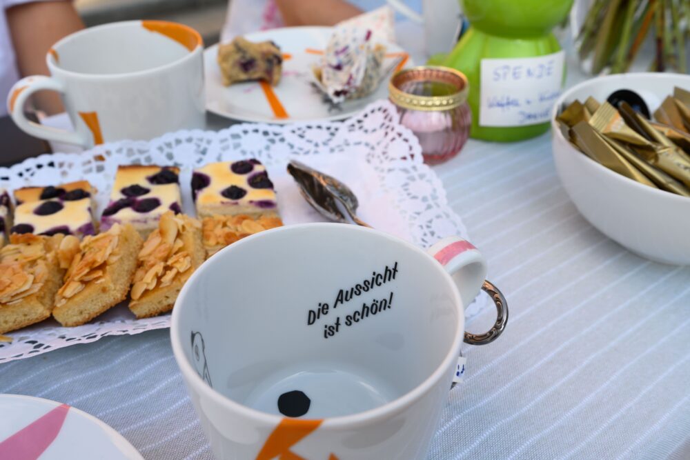 Das Bild zeigt einen gedeckten Tisch. Es ist eine Tasse zu sehen auf der steht "die Aussicht ist schön!". Dahinter stehen Kuchenstückchen.