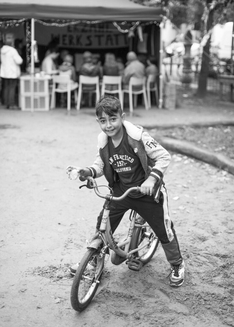 Ein Junge sitzt auf seinem Fahrradsattel und steht mit dem Rad. Er steht zentral im Bild und schaut in die Kamera.