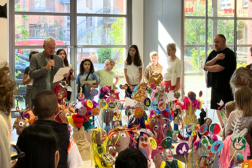 In der Galerie im Atelierhaus Roter Hahn wird eine Ausstellung eröffnet. Kinder und Erwachsene hören einem Redner zu. In der Mitte zwischen den Personen sind Skulpturen und Plastiken zu sehen.