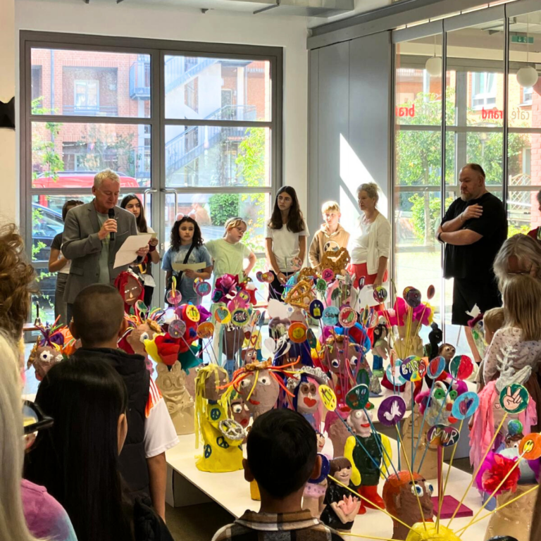 In der Galerie im Atelierhaus Roter Hahn wird eine Ausstellung eröffnet. Kinder und Erwachsene hören einem Redner zu. In der Mitte zwischen den Personen sind Skulpturen und Plastiken zu sehen.