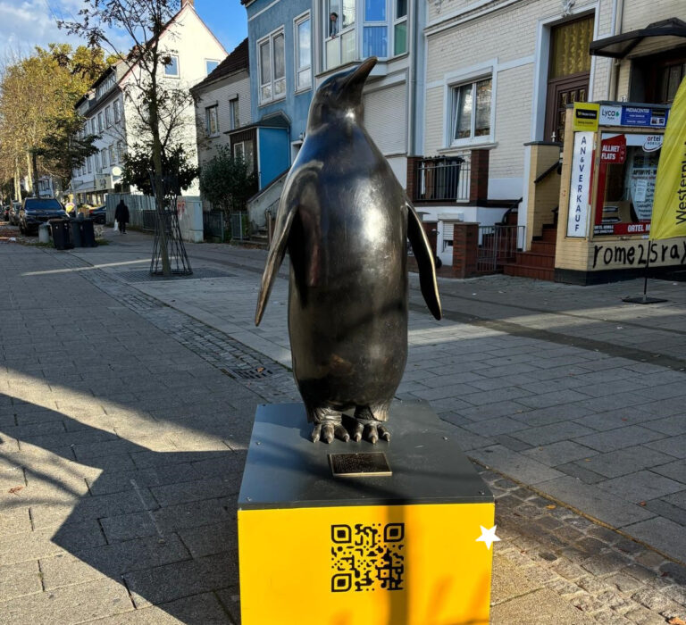 Eine Bronzeskulptur in Form eines Pinguins steht auf einem gelben Sockel. Auf dem Sockel ist ein QR-Code.