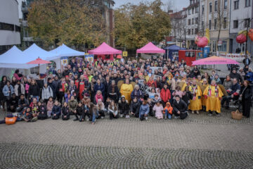 Auf einem großen Platz sind viele Menschen, darunter auch viele Kinder und Menschen in Kostümen zusammengekommen um fotografiert zu werden. Sie lächeln in die Kamera.