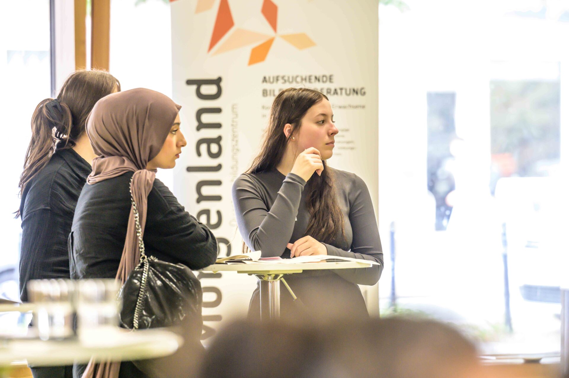 Drei Personen stehen an einem Stehtisch und schauen alle in eine Richtung. eine Person lehnt ihren Ellenbogen auf den Tisch. Im Hintergrund ist ein Aufsteller zu sehen.