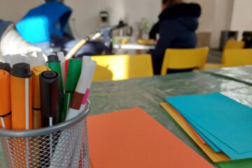 Ein Metallbehälter mit farbigen Filzstiften steht im Vordergrund auf einem Tisch, der mit einer durchsichtigen Plastikdecke bedeckt ist. Daneben liegen bunte Blätter Papier in Orange, Blau und Grün. Im Hintergrund sind zwei Personen an einem Tisch mit gelben Stühlen unscharf zu erkennen, während Sonnenlicht den Raum teilweise erhellt.