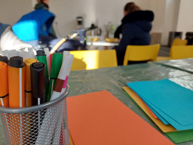 Ein Metallbehälter mit farbigen Filzstiften steht im Vordergrund auf einem Tisch, der mit einer durchsichtigen Plastikdecke bedeckt ist. Daneben liegen bunte Blätter Papier in Orange, Blau und Grün. Im Hintergrund sind zwei Personen an einem Tisch mit gelben Stühlen unscharf zu erkennen, während Sonnenlicht den Raum teilweise erhellt.