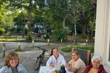 Ein gedeckter Tisch steht draußen neben einem Haus. Vier Personen sitzen am Tisch und schauen in die Kamera. Im Hintergrund ist eine Reihe Bäume zu erkennen.