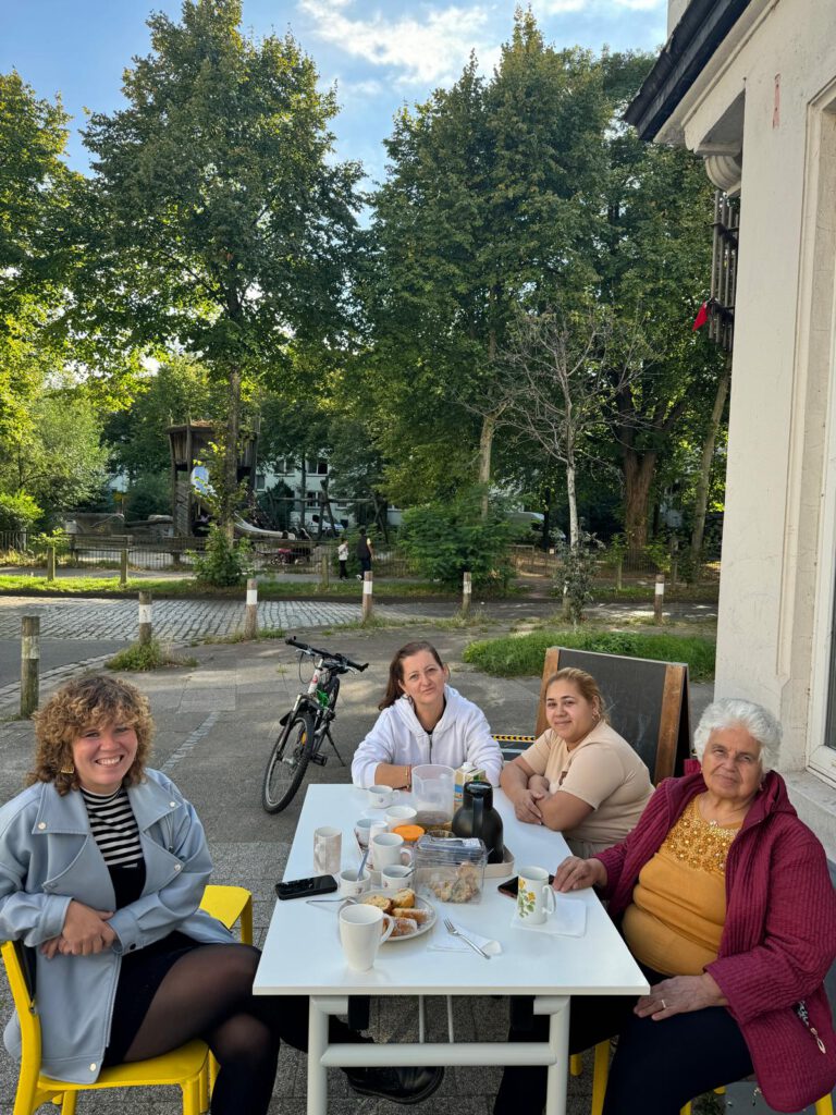 Ein gedeckter Tisch steht draußen neben einem Haus. Vier Personen sitzen am Tisch und schauen in die Kamera. Im Hintergrund ist eine Reihe Bäume zu erkennen.