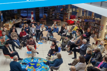 Ein Foto von oben auf eine Gruppe sitzender Menschen in einem großen Raum. Eine Person spricht in ein Mikrofon, die anderen hören zu.