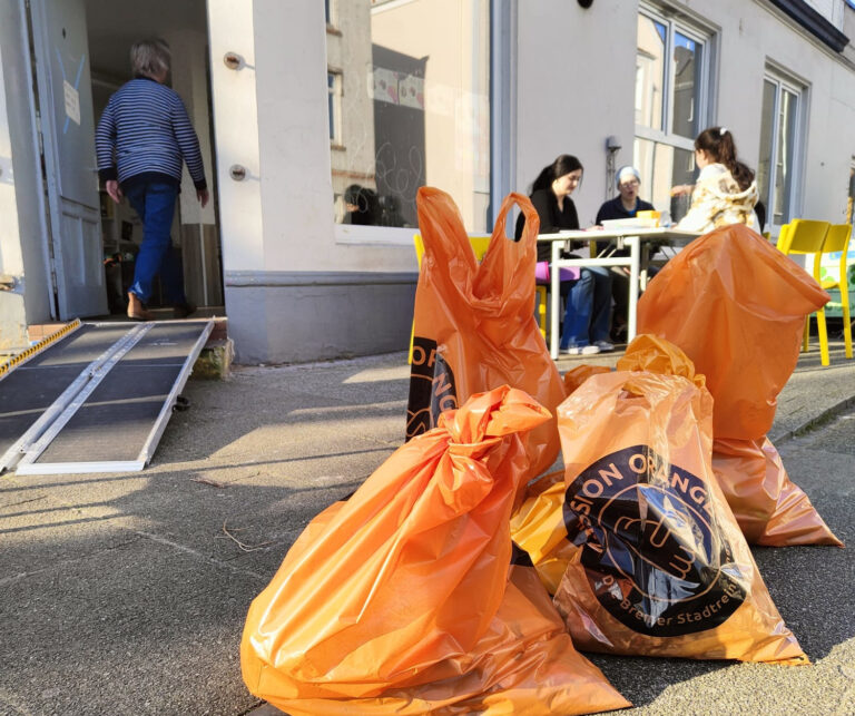 Gefüllte orangene Müllsäcke liegen auf einem Vorplatz vor einem Nachbarschaftstreff. Im Hintergrund geht eine Person ins Haus, rechts davon sitzen drei Personen.