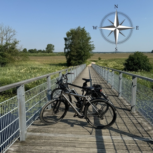 zwei Fahrräder stehen nebeneinander auf einer Rad- und Fußgängerbrücke. Weiter hinten ist ein Wanderweg zu sehen zwischen Feld und Wiesen.