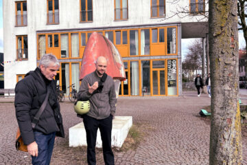 Zwei Personen sind ins Gespräch vertieft. sie schauen einen Baum auf einen Platz an. Im Hintergrund ist ein großes Gebäude zu sehen und vorbeigehende Menschen.