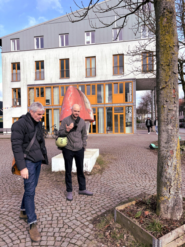 Zwei Personen sind ins Gespräch vertieft. sie schauen einen Baum auf einen Platz an. Im Hintergrund ist ein großes Gebäude zu sehen und vorbeigehende Menschen.