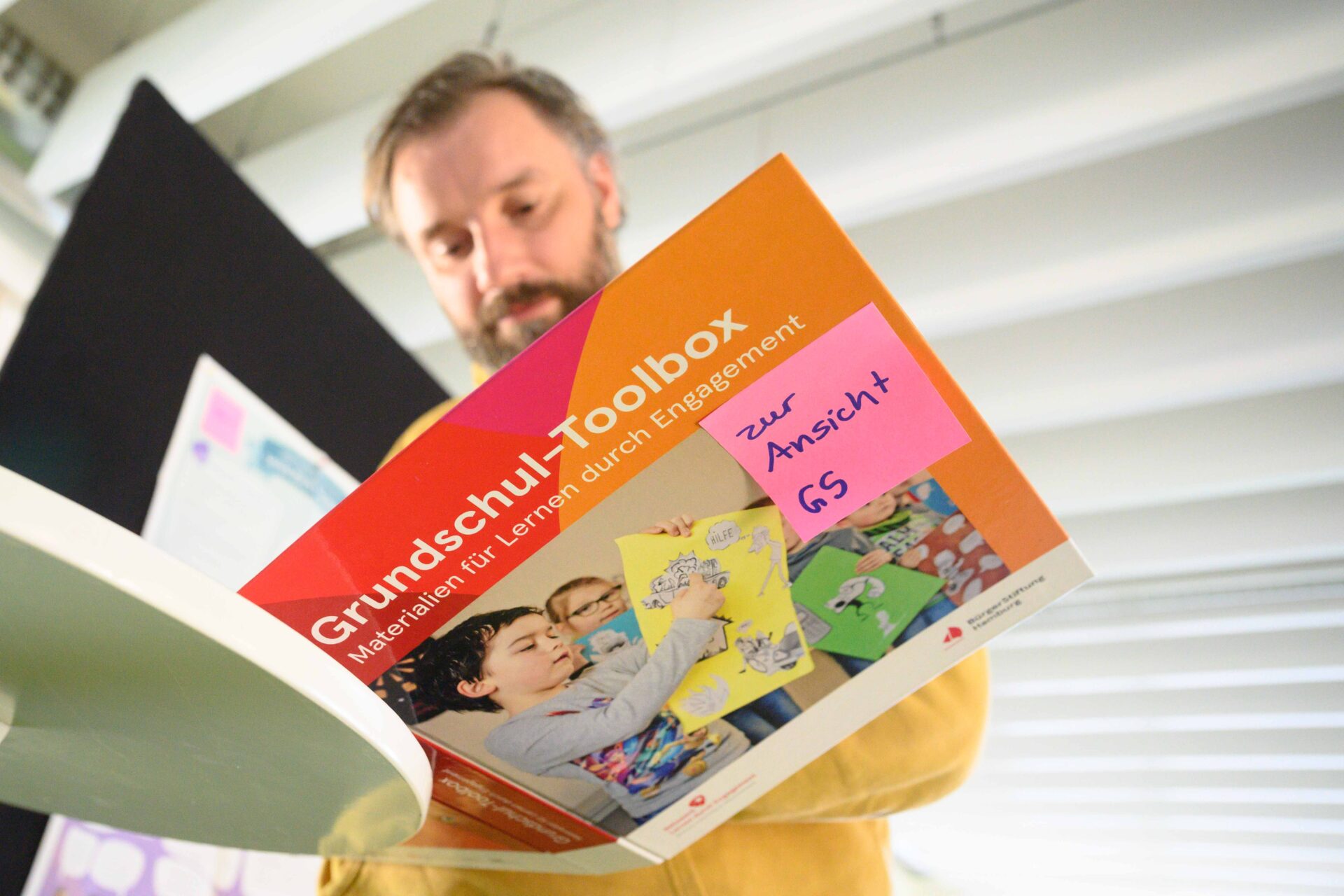 Ein Blick von unten nach oben: Eine Person hält eine Broschüre vor sich aufgeschlagern. Darauf steht: "Grundschul-Toolbox. Materialien für Lernen durch Engagement. Ein Post-it klebt auf der Broschüre, darauf steht "Zur Ansicht GS". Auf der Broschüse ist auch ein Foto mit Kindern, die etwas auf einem Pasier zeigen.