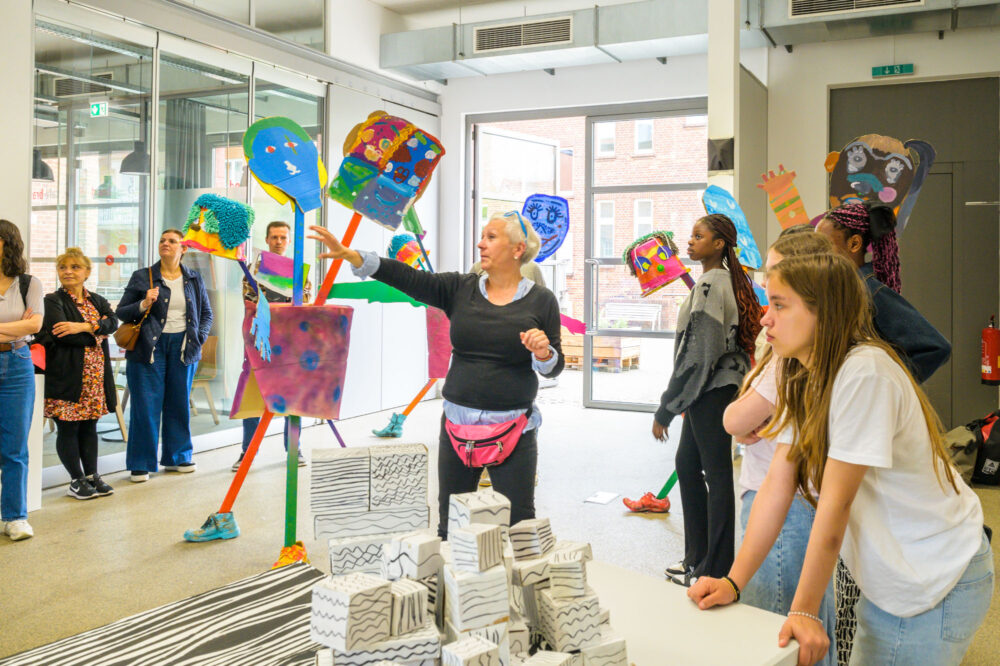In einer Ausstellung mit Kunstwerken im Raum erklärt eine Person gestikulierend das zu Sehende. An den Seiten stehen Personen und hören und schauen zu.