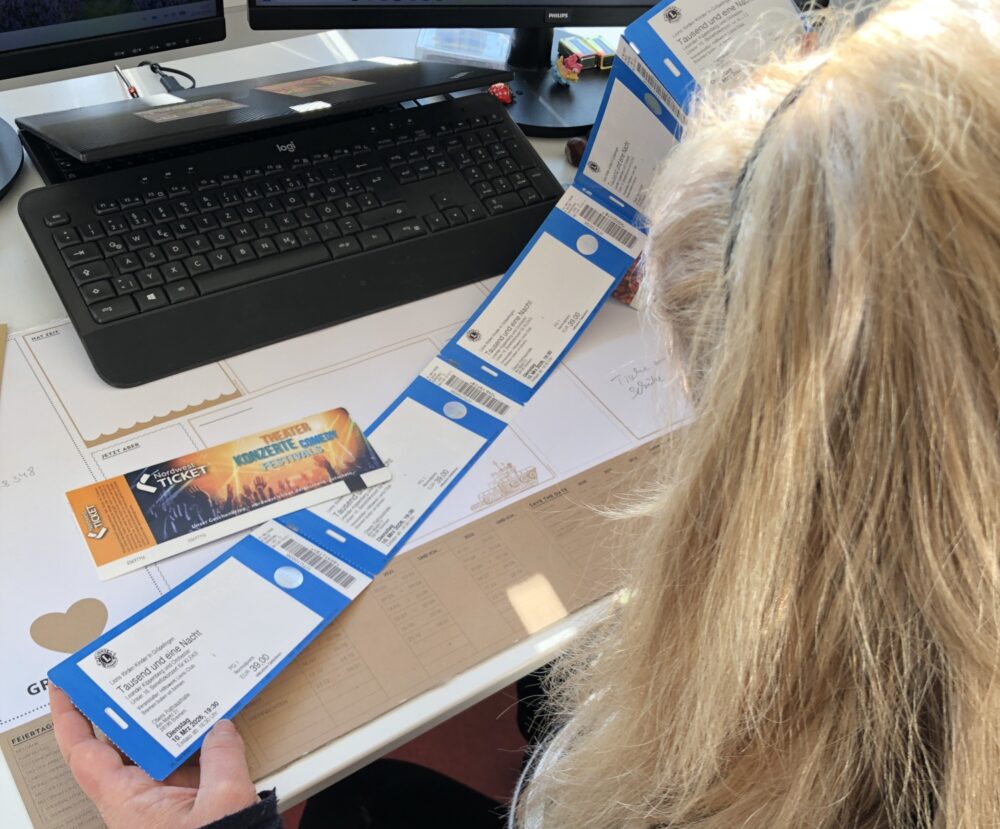Über die Schulter geblickt sehen wir wie eine sitzende Person mit langem Haar Tickets in ihren Händen hält. Ein Laptop und ein Bildschirm sind ebenfalls zu sehen.
