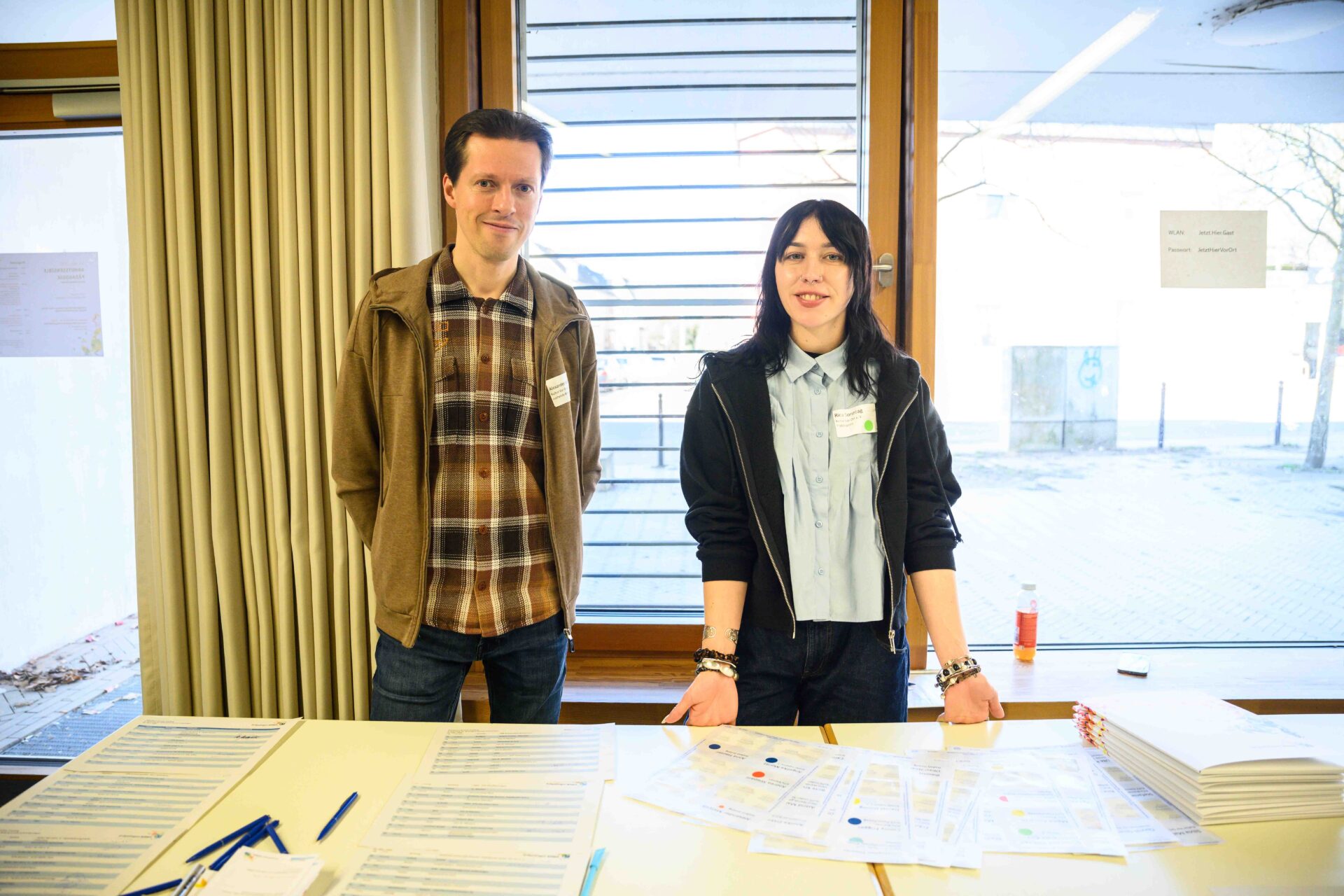 Zwei Personen stehen hinter einem Tisch mit Anmeldebögen. Eine Person lehnt sich mit beiden Händen gegen die Tischkante.