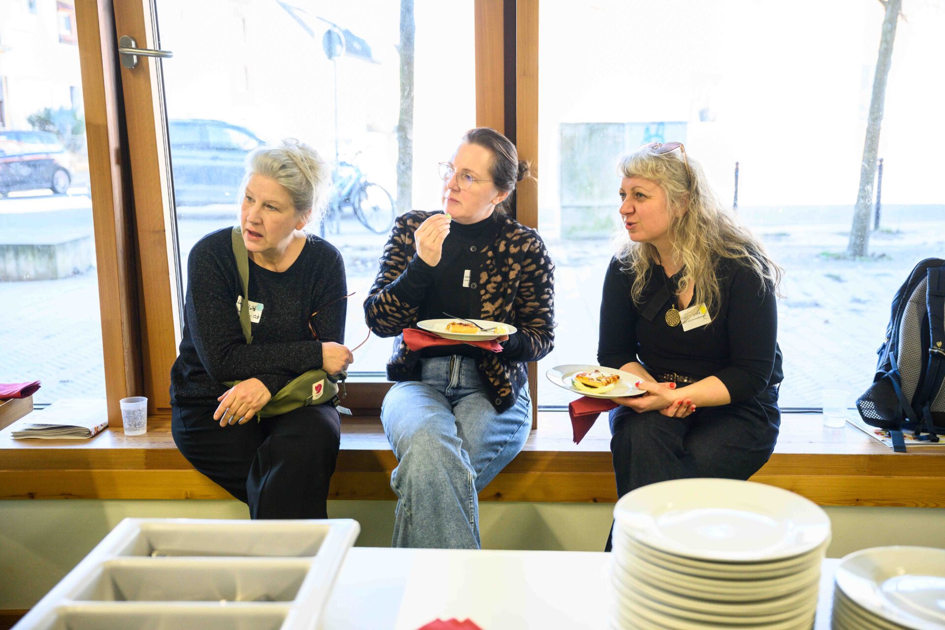 Drei Personen sitzen auf einer Fensterbank teilweise mit etwas zu Essen in der Hand. Sie schauen seitlich nachrechts und scheinen zu sprechen.