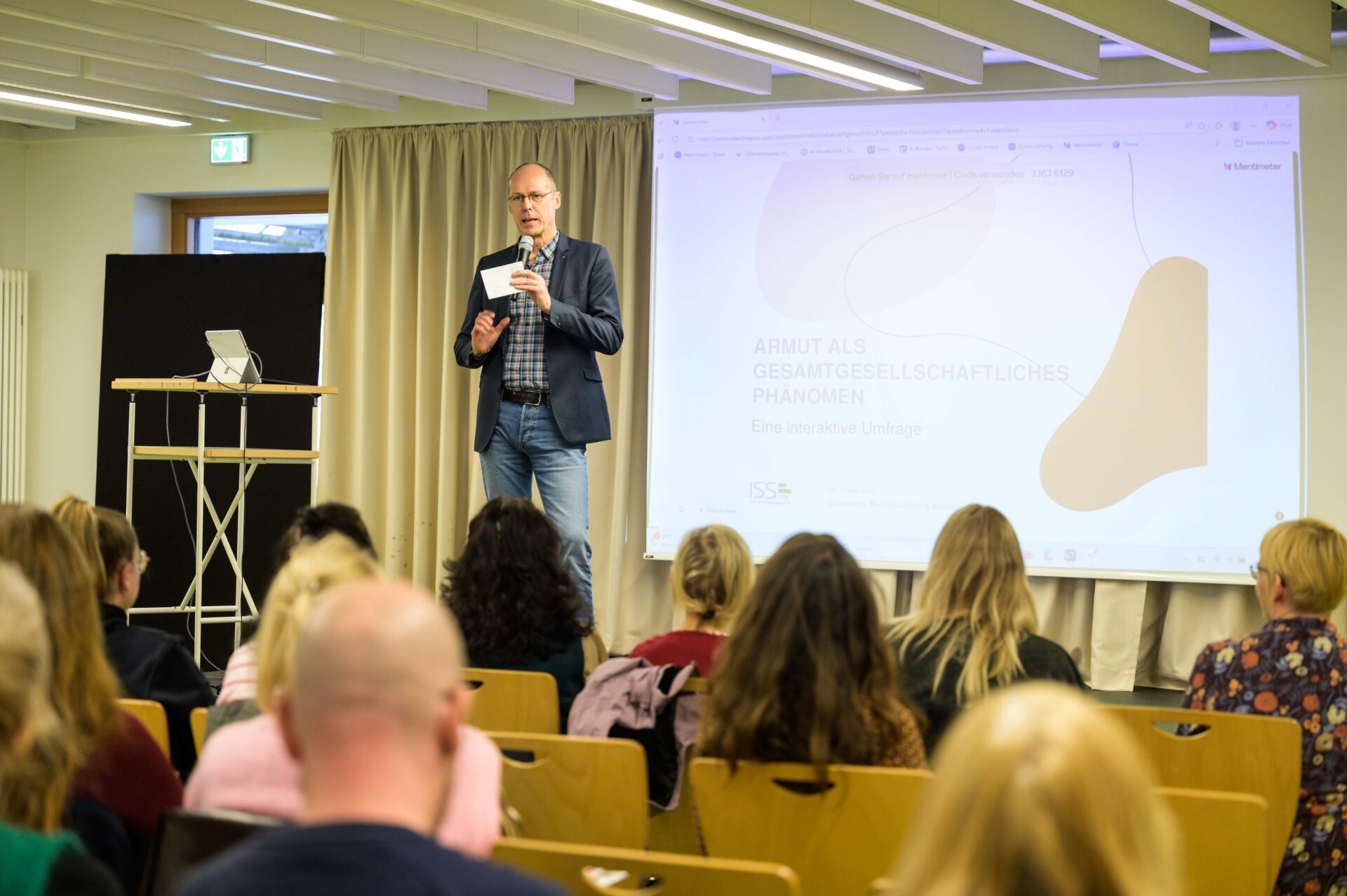 Auf einer Bühne steht eine Person und hält ein Mikrofon und Zettel in der einen Hand, gestikuliert mit der anderen und redet vor sitzendem Publikum.