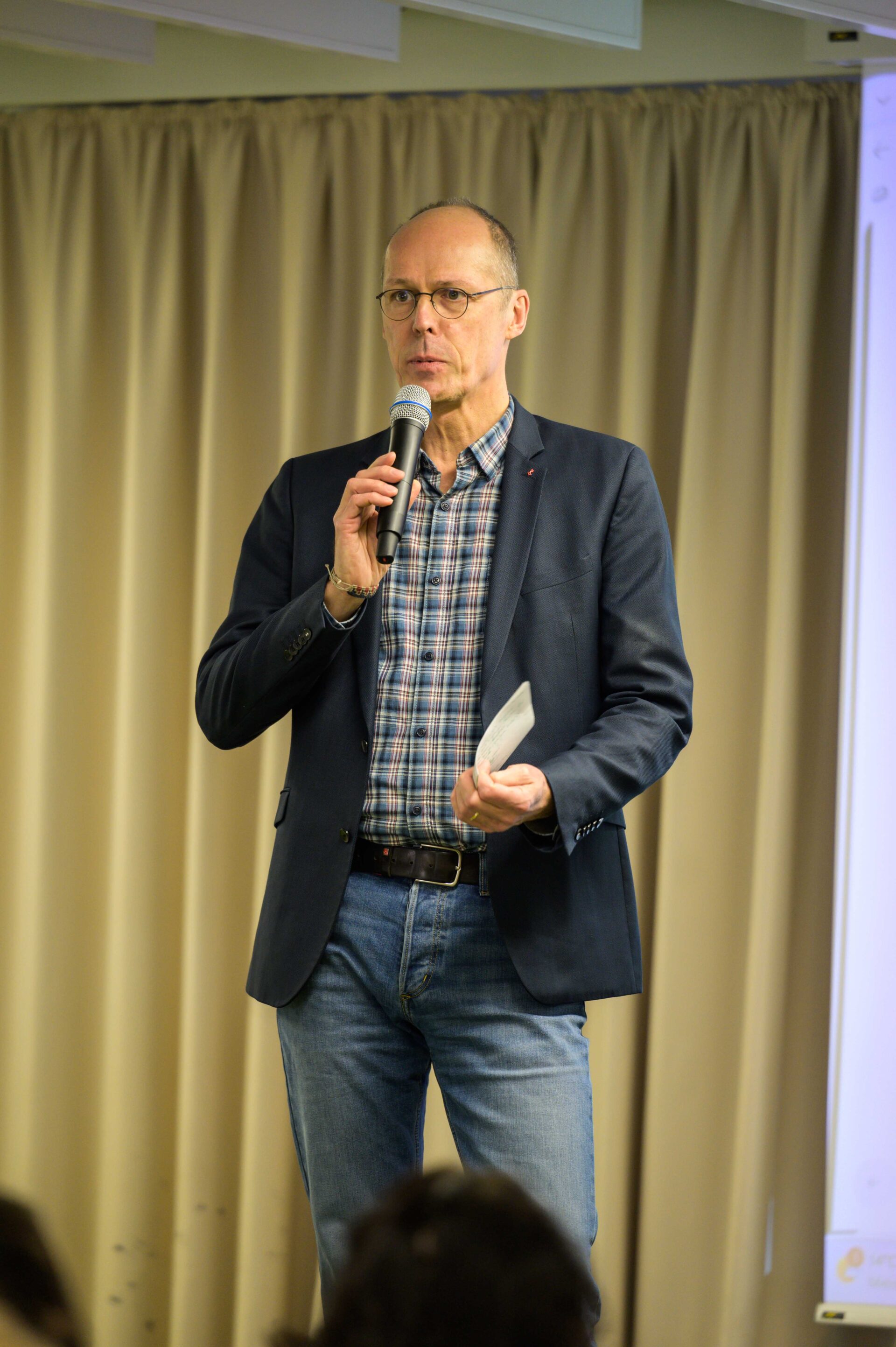 Eine Person steht vor einem Vorhang und hält ein Mikrofon und Zettel in der einen Hand, gestikuliert mit der anderen und redet