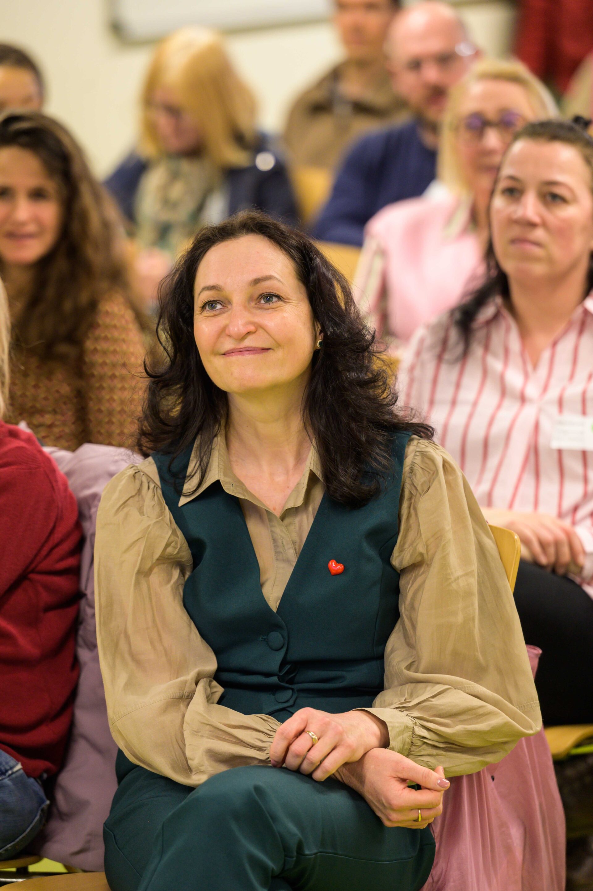 Eine Person mit langen dunklen Haaren sitzt zusammen mit anderen Personen in Stuhlreihen und lächelt nach vorne.