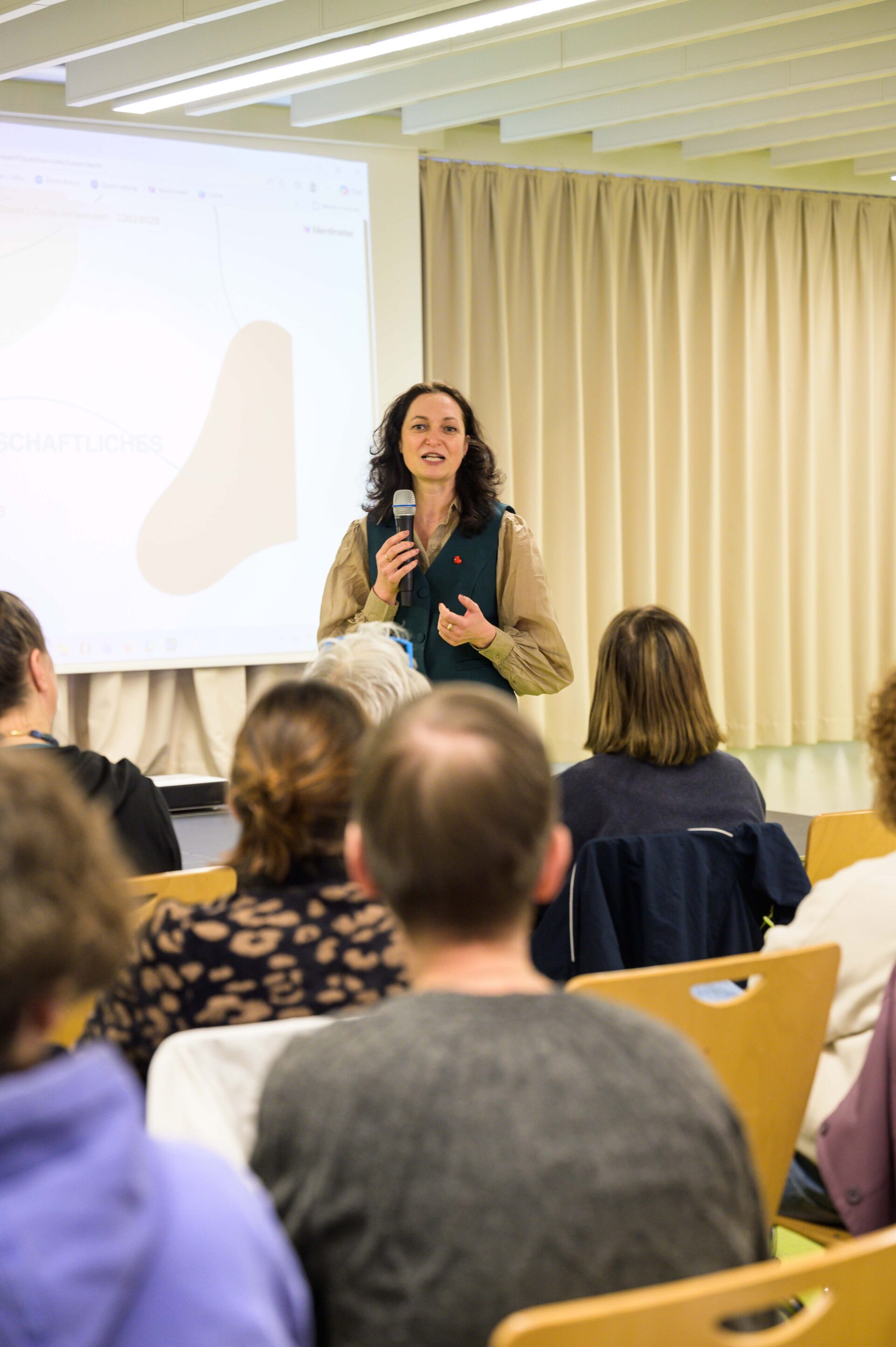 Auf einer Bühne befindet sich im Hintergrund eine Leindwand, auf die etwas projeziert wird. Vor der Bühne steht eine Person mit langen Haaren und Mikrofon in der einen Hand, während sie mit der anderen Hand gestikuliert. Sie spricht dabei. Vor der Bühne sitzen auf Stühlen Personen die zu der Person auf der Bühne blicken und zuhören.