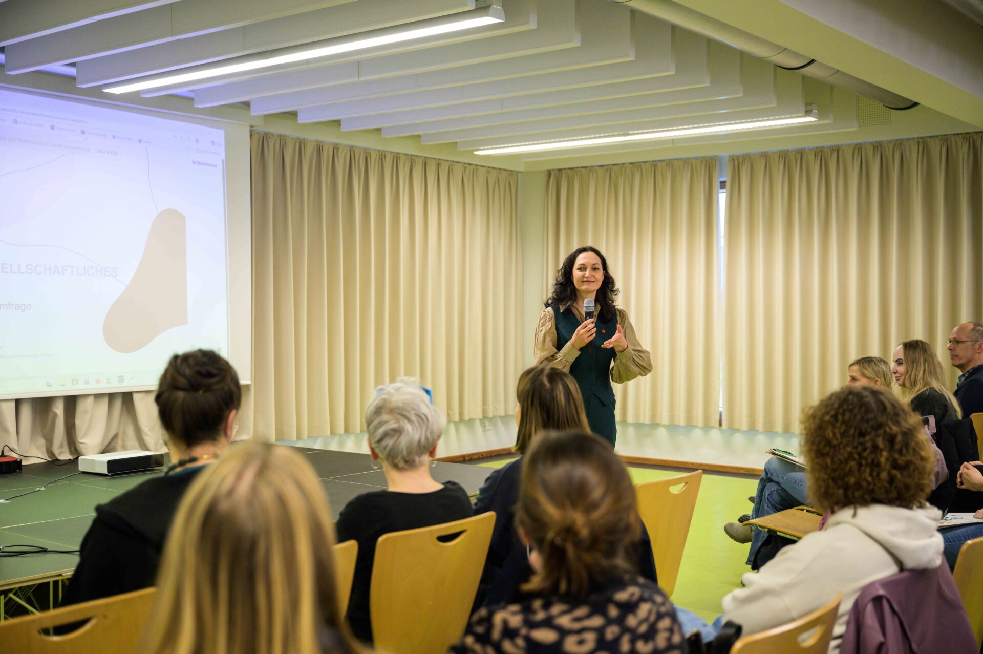 Auf einer Bühne befindet sich im Hintergrund eine Leindwand, auf die etwas projeziert wird. Vor der Bühne steht eine Person mit langen Haaren und Mikrofon in der einen Hand, während sie mit der anderen Hand gestikuliert. Sie spricht dabei. Vor der Bühne sitzen auf Stühlen Personen die zu der Person auf der Bühne blicken und zuhören.