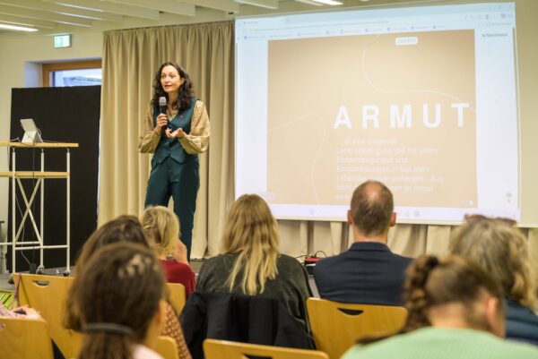 Auf einer Bühne befindet sich im Hintergrund eine Leindwand, auf die etwas projeziert wird. Davor steht eine Person mit langen Haaren und Mikrofon in der einen Hand, während sie mit der anderen Hand gestikuliert. Sie spricht dabei. Vor der Bühne sitzen auf Stühlen Personen die zu der Person auf der Bühne blicken und zuhören.