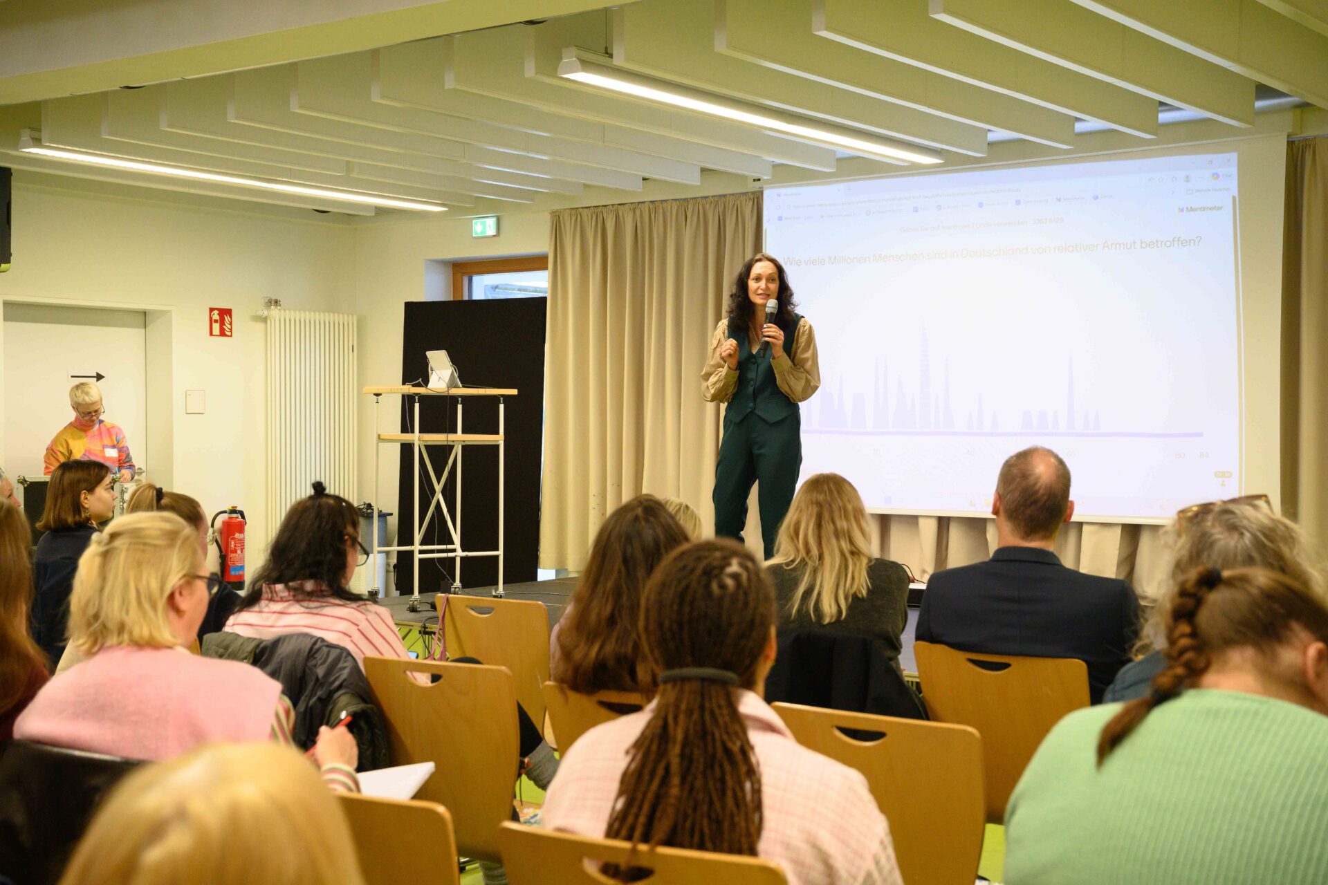 Auf einer Bühne befindet sich im Hintergrund eine Leindwand, auf die etwas projeziert wird. Davor steht eine Person mit langen Haaren und Mikrofon in der einen Hand, während sie mit der anderen Hand gestikuliert. Sie spricht dabei. Vor der Bühne sitzen auf Stühlen Personen die zu der Person auf der Bühne blicken und zuhören.