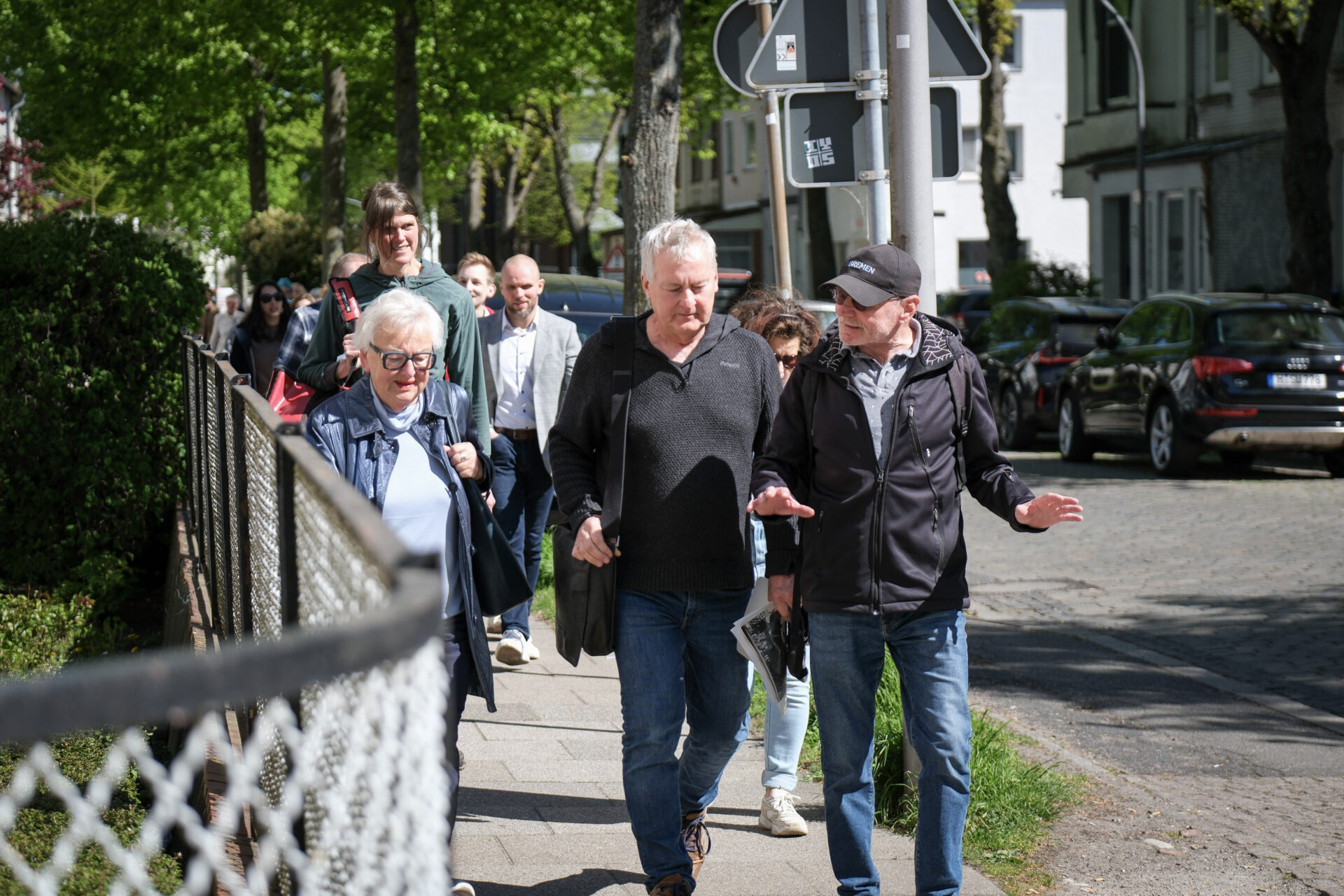 Eine Gruppe von Menschen bewegt sich entlang einer Straße. Die ersten beiden sind ins Gespräch vertieft.