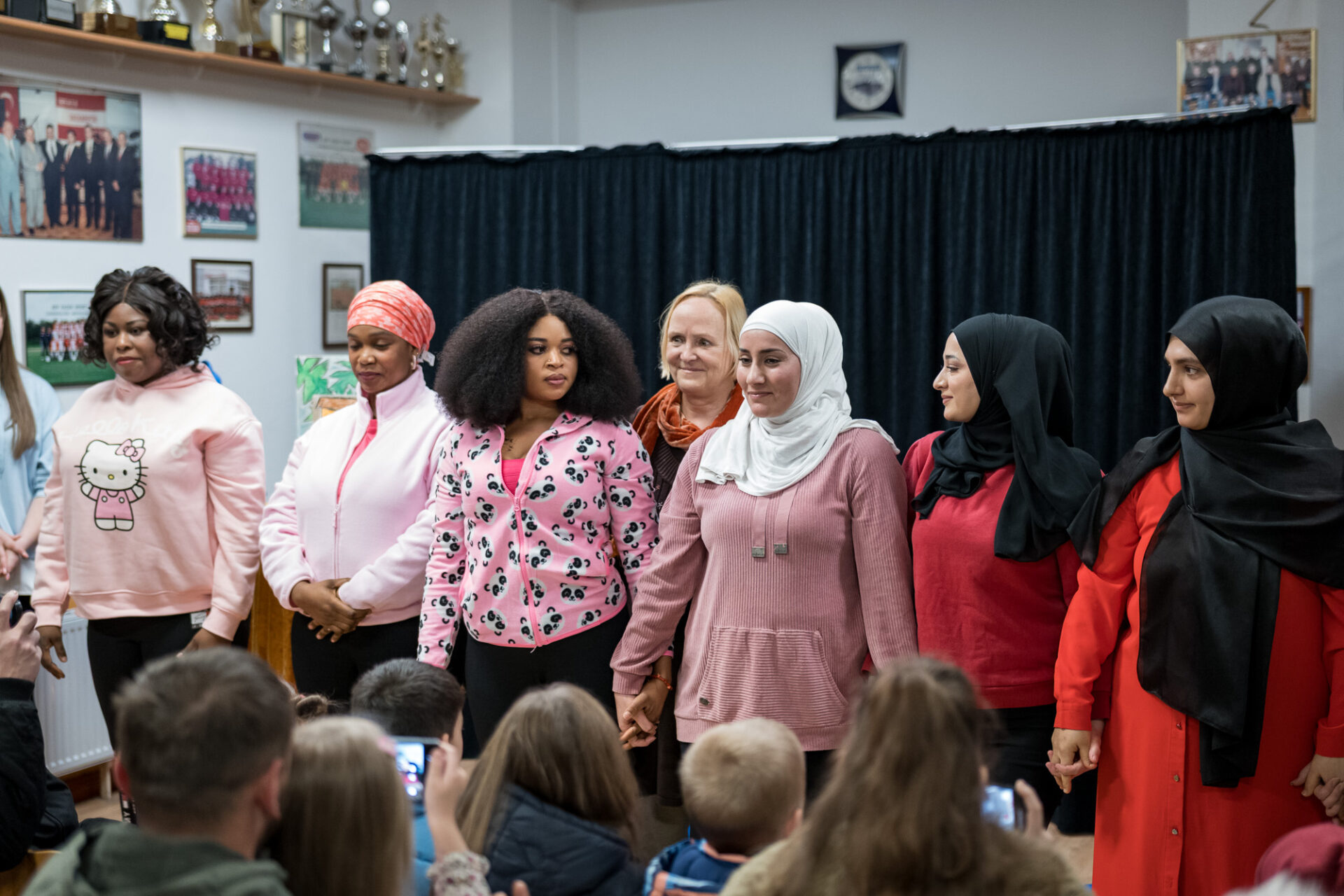 Sieben Personen stehen händchenhaltend nebeneinander. Hinter Ihnen ist ein schwarzer Bühnenvorhang, vor ihnen sitzt Publikum.