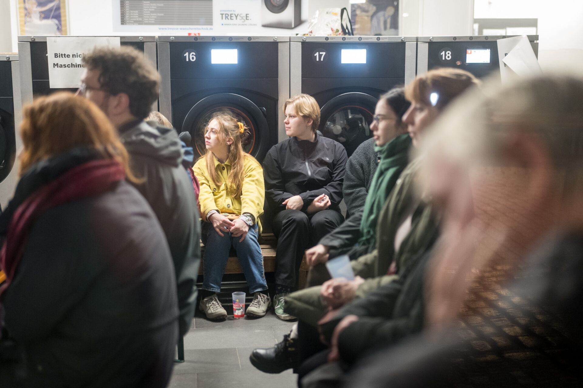 Im Waschcenter sitzen Personen auf Bänken und schauen alle in eine Richtung. Im Hintergrund sind Waschmaschinen.