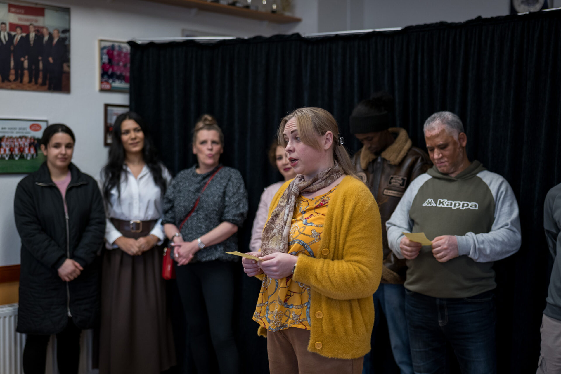 Mehrere Personen stehen vor einem schwarzen Bühnenvorhang, eine spricht.