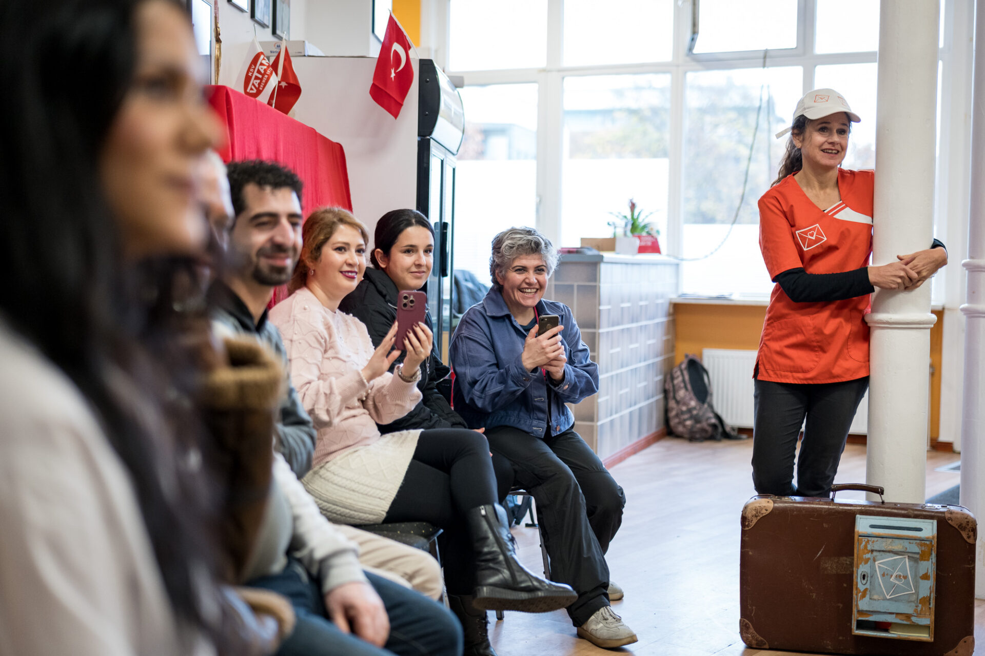 Mehrere Personen sitzen auf Stühlen und lächeln. Eine Person steht an einer Säule, davor steht ein Koffer.