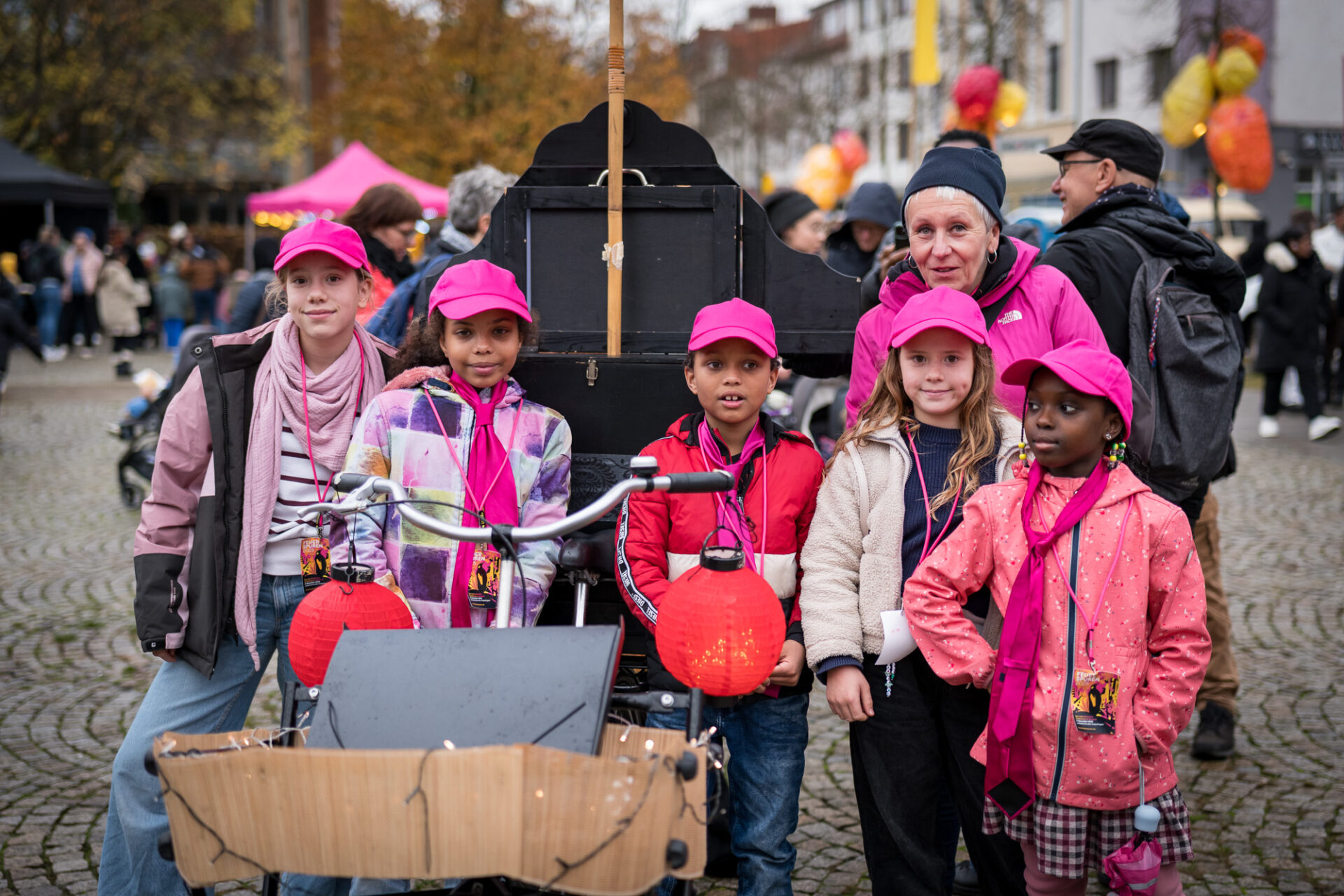 Fünf Kinder in pinker Kleidung und eine Erwachsene schauen in die Kamera. Im Hintergrund ist ein Platz mit Menschen zu sehen und eine schwarze Kiste.