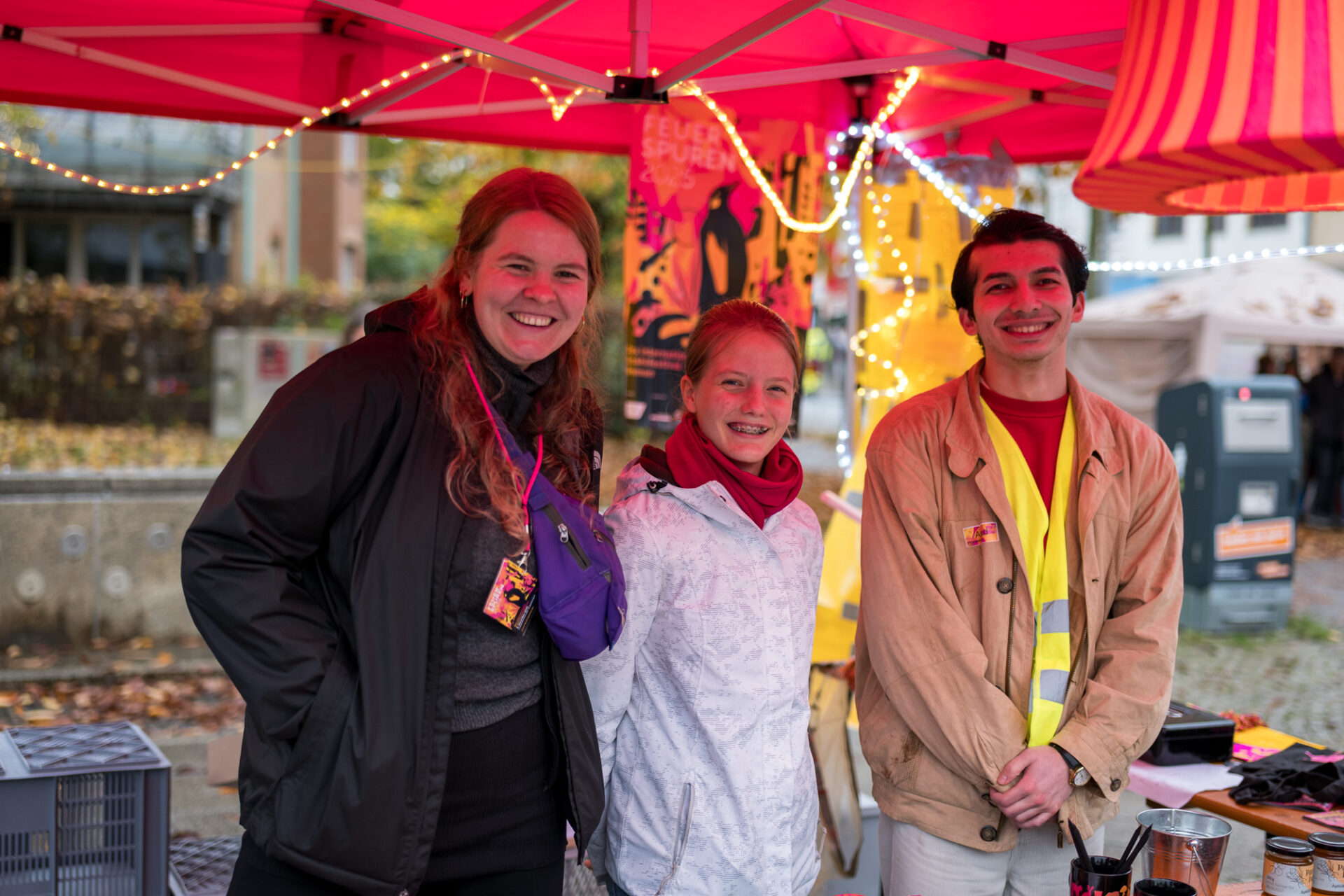 Unter einem pinken Pavillon stehen drei Personen und lächeln freundlich in die Kamera. Im Hintergrund ist ein Plakat von dem internationalen Erzählfestival Feuerspuren. Im Pavillon hängt ein Lichterschlauch und ein Lampenschirm.