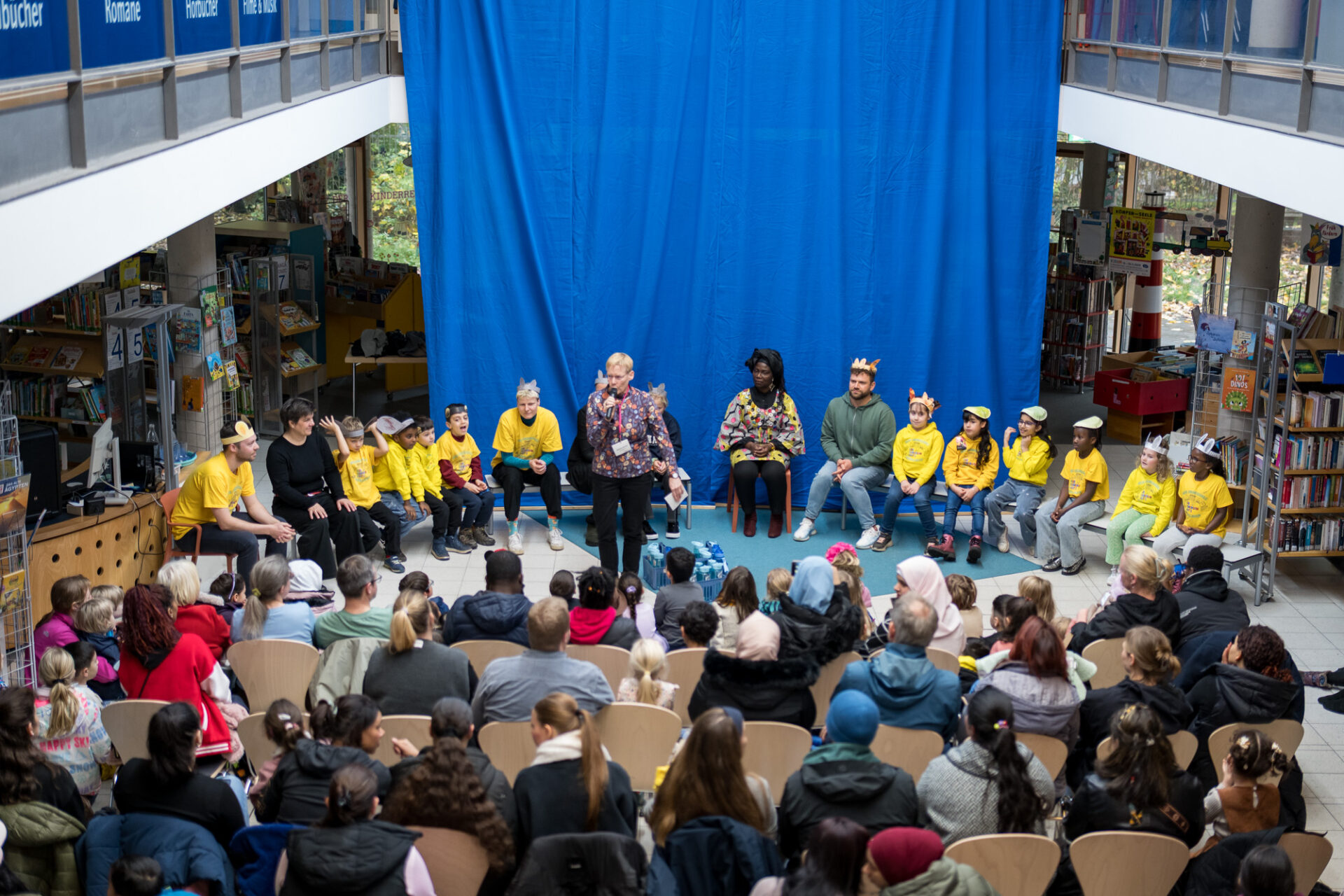 Ein Blick aus der Vogelperspektive auf einen großen Raum. Auf der Bühne sitzen viele Kinder in gelben T-Shirts und ein paar Erwachsene dazwischen. Eine Person in buntem Hemd steht und spricht in ein Mikrofon. Davor sind Stuhlreihen. Hier sitzen viele Menschen und schauen Richtung Bühne.