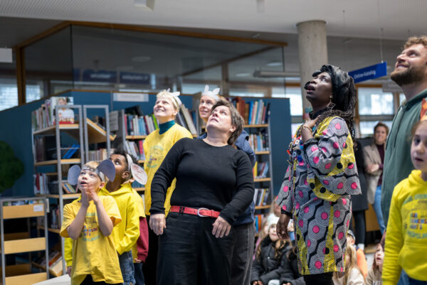 Mehrere bunt gekleidete Personen unterschiedlichen Alters schauen ausdrucksstark nach oben.