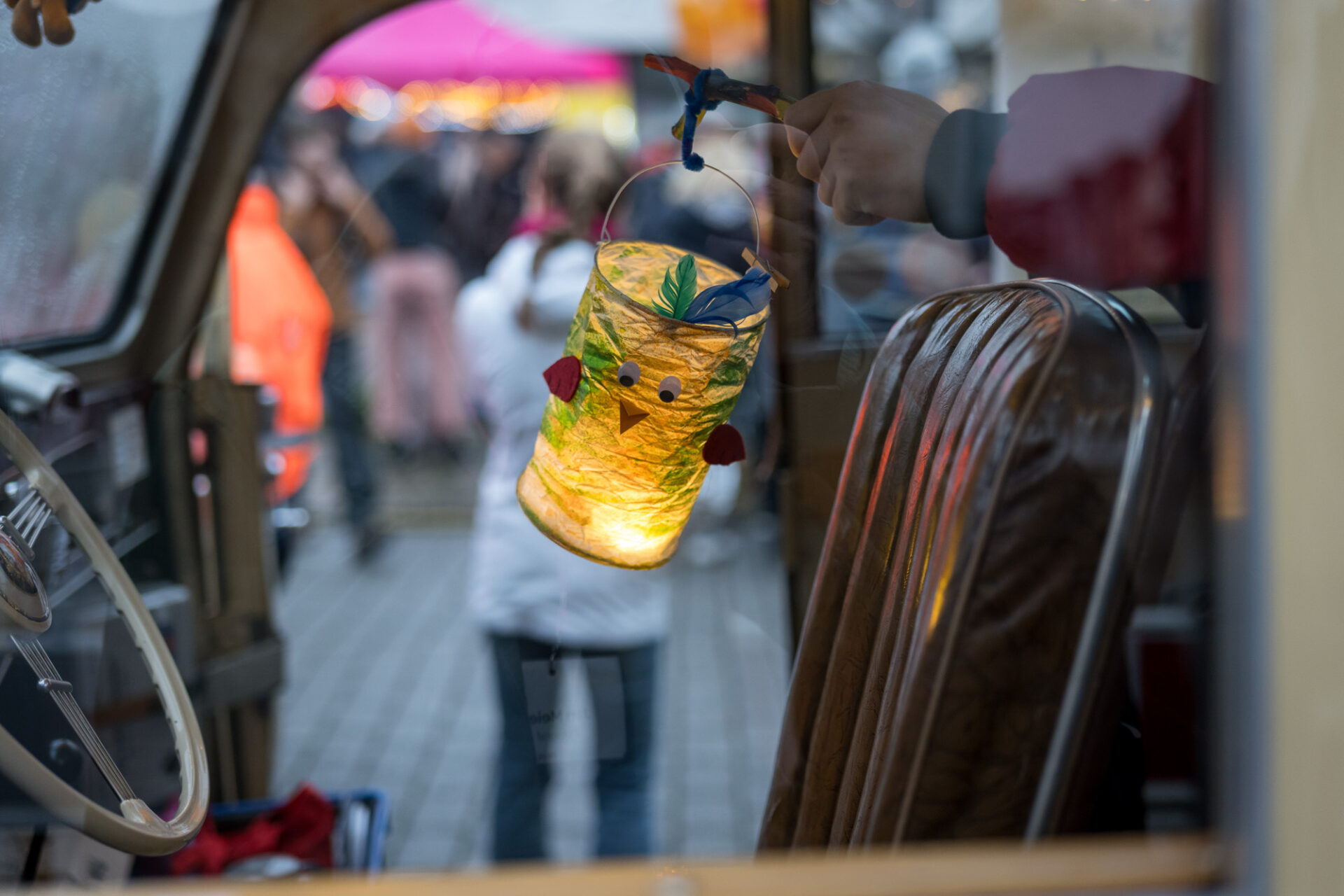 Eine Hand hält eine Laterne mit Vogelgesicht und Federn. Im Bild befindet sich ein Lenkrad und ein Fahrersitz, die Autotür ist geöffnet. Im Hintergrund befinden sich Personen und ein pinker Baldachin.