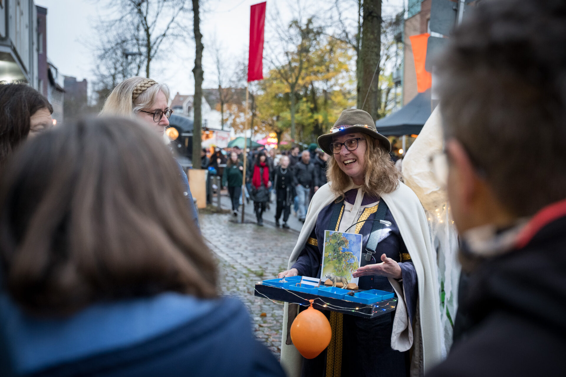 Eine bunt gekleidete Person mit Bauchladen gestikuliert mit einer Hand und spricht mit Menschen auf der Straße.