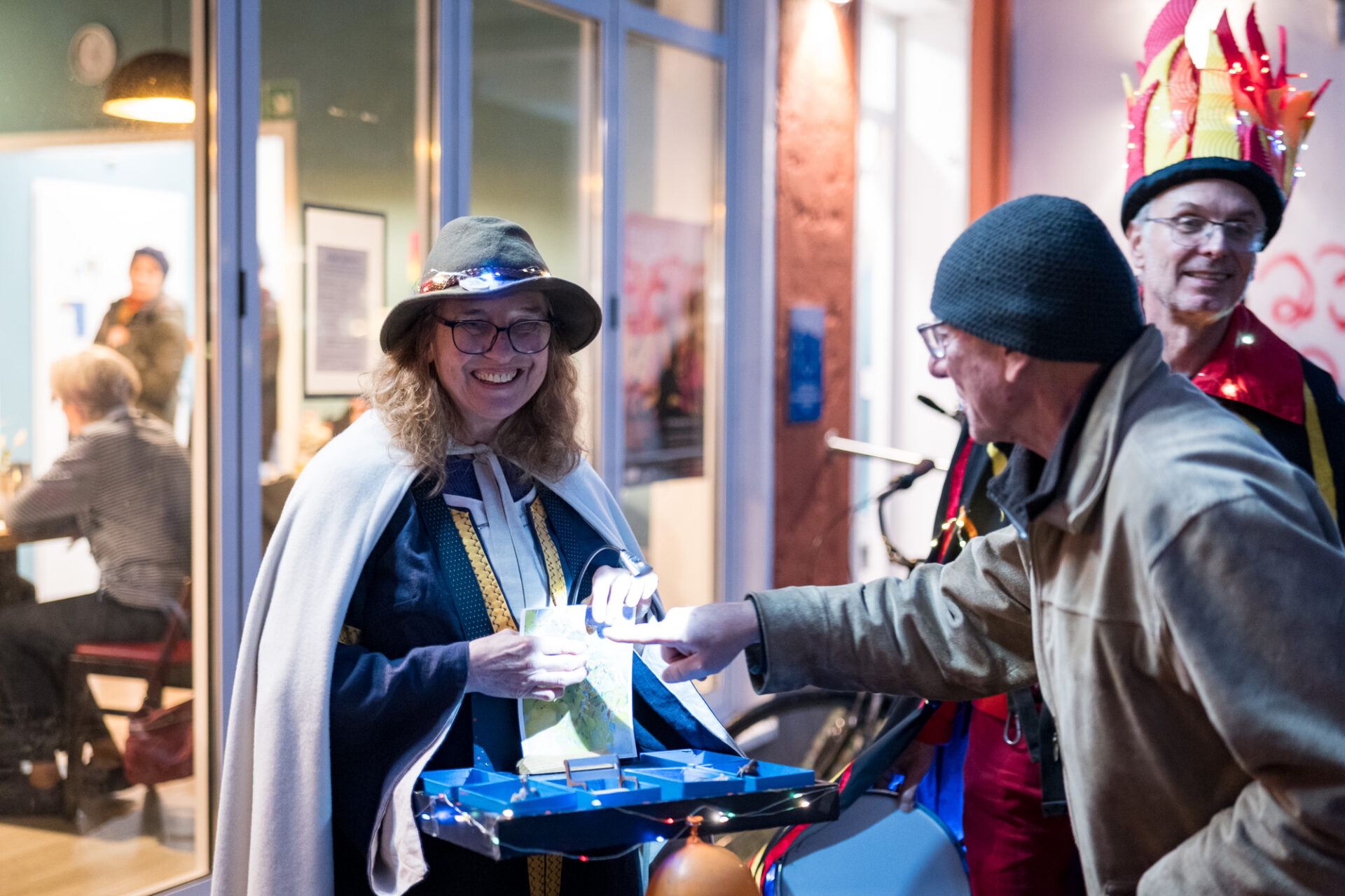 Eine lustig gekleidete Person mit Hut hat einen Bauchladen vor der Brust. Eine Person steht davor und zeigt mit ausgestrecktem Finger auf etwas beim Bauchladen. Eine weitere Person mit bunter Kleidung und Hut lugt hinter dieser Person hervor und lächelt in die Kamera.