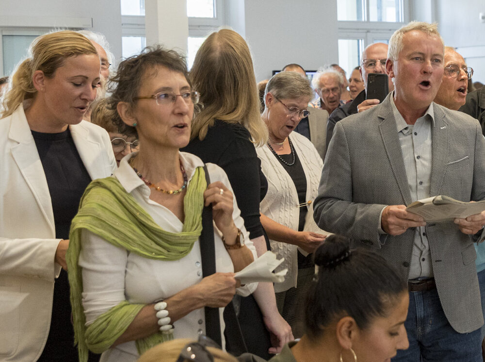 In einem Raum stehen mehrere festlich gekleidete Personen eng beieinander und singen.