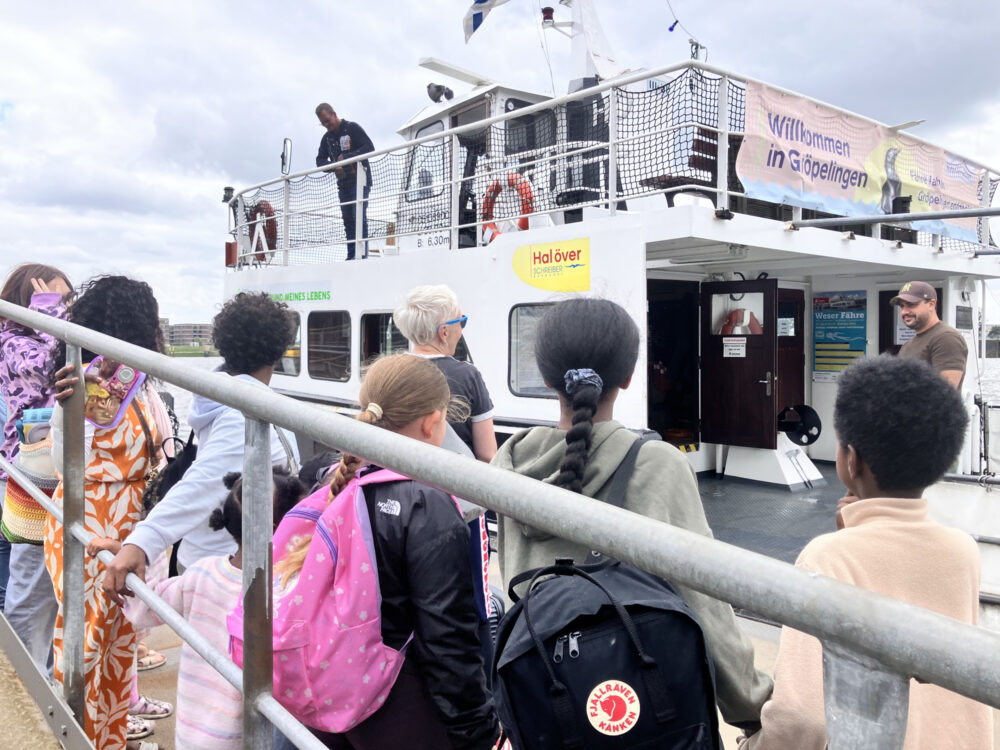 An einem Schwimmanleger warten viele Kinder auf eine Fähre, die gerade angelegt hat. An Deck stehen Schiffführer und Lotse.