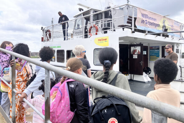 An einem Schwimmanleger warten viele Kinder auf eine Fähre, die gerade angelegt hat. An Deck stehen Schiffführer und Lotse.