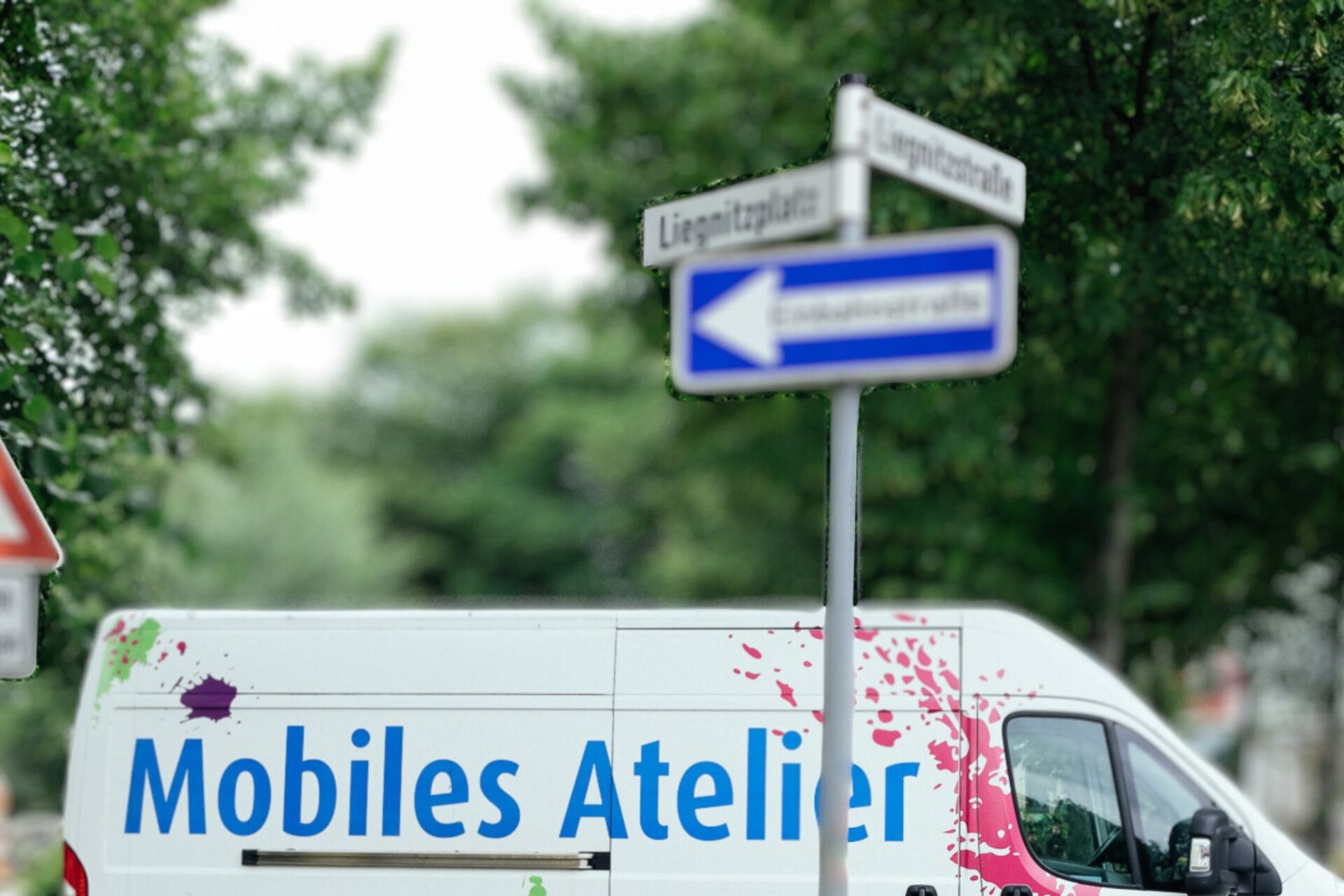 Ein weißer Transporter trägt die Aufschrift: Mobiles Atelier und Farbklekse alsa Dekoration. Davor steht ein Straßenschild an dem auch ein Einbahnstraßenschild befestigt ist. Im hintergrund sind Bäume.