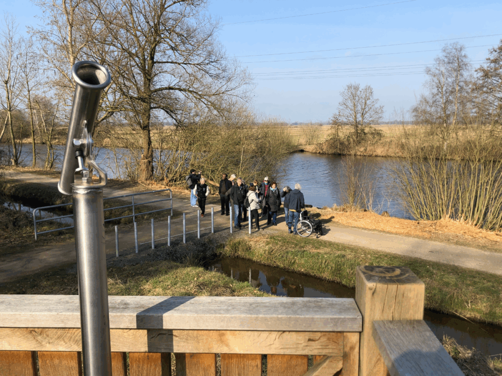 Im Vordergrund ist eine Aussichtsrohr auf einem Holzgeländer zu sehen. Im Hintergrund spaziert eine Gruppe von Menschen entlang eines Weges am Flussufer. Einige Personen sind zu Fuß unterwegs, während eine andere Person im Rollstuhl sitzt. Der Himmel ist klar und es sind Bäume und der Fluss zu sehen.